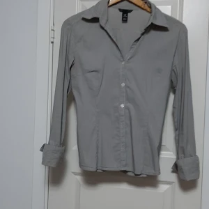 Gråvit randig skjorta H&M strl 38 - Klassisk skjorta från H&M i storlek 38 med smala grå och vita ränder. Skjortan har lång ärm, knappar framtill och en snygg krage. Tillverkad i bomull, perfekt för en clean och stilren look.