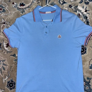 Blå Moncler pikétröja med logga - Snygg ljusblå pikétröja från Moncler med klassisk krage och knappar framtill. Tröjan har Moncler-logga på bröstet och röd, vit och marinblå rand på krage och ärmslut. Perfekt för en stilren och sportig look.