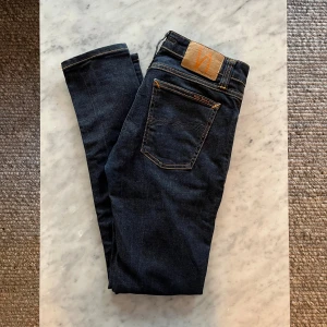 Nudie Jeans  - Nudie Jeans 28/30 – Klassisk mörk denim Snygga och bekväma Nudie Jeans i storlek 28/30. Mörkblå tvätt med subtila slitningar och den ikoniska Nudie-sömmen på bakfickan. Tillverkade i högkvalitativ denim med perfekt passform – både stilrena och hållbara. Perfekta för både vardag och fest! ::;::;;::;;;::;::;;:::;:::;:::;:::;:::;;;::;;;:::;;:::;;;:;;;;;;;;;;;;;::;;::::::::;;:::- Mörkblå jeans - Brun läderlapp - Varumärket Nudie Jeans - Bakficka - Dekorativa sömmar