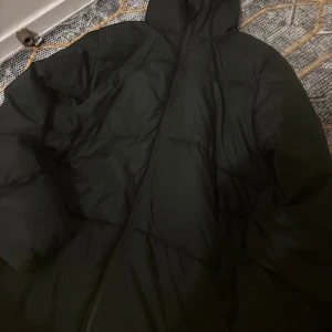 Svart pufferjacka med dragkedja - Säljer en svart pufferjacka med hög krage och hel dragkedja framtill. Jackan är vadderad och har en clean, oversized look. Perfekt för kalla dagar och riktigt skön att bära. Fodrad insida och praktiska fickor. Passar dig som gillar streetstyle.