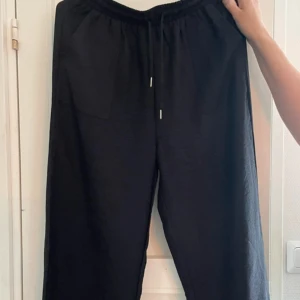 Svarta vida byxor  - Svarta byxor med bred passform och resår i midjan samt snörning framtill. Byxorna har två stora fickor framtill och är tillverkade i ett mjukt tyg som ger en avslappnad look. Perfekta för en chill och stilren outfit.