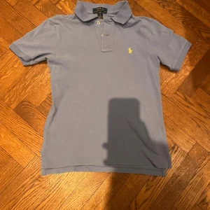 Ljusblå pikétröja från Ralph Lauren - Säljer en klassisk ljusblå pikétröja från Ralph Lauren med gul broderad logga på bröstet. Barnstorlek S(8) pojke. Tröjan har korta ärmar, krage och knäppning framtill. Perfekt för en clean och stilren look.