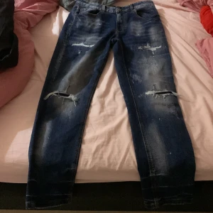 Blå slitna jeans Supply & Demand - Säljer ett par blå jeans från Supply & Demand med slitna detaljer och hål på knäna. Jeansen har klassisk femficksdesign, normal passform och är tillverkade i denim. Perfekt för dig som gillar en edgy streetstil.