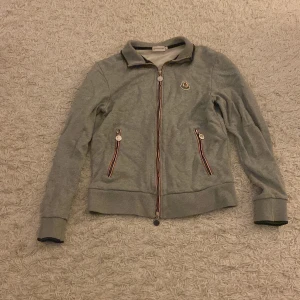 Grå Moncler kofta med dragkedja - Säljer en grå kofta från Moncler med klassisk logga på bröstet. Tröjan har hel dragkedja framtill och två fickor med snygga rödblå detaljer. Mjuk och skön bomullskvalitet, perfekt för lager-på-lager. Långärmad och med ribbade muddar för extra komfort.