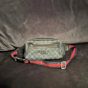 Svart magväska  - Snygg svart magväska med justerbart band i rött och marinblått. Väskan har dragkedja framtill och detaljer i skinn. Det finns vissa bekymmer med väskan, skriv för mer info.