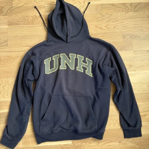 Mörkblå Champion UNH hoodie M - Mörkblå hoodie från Champion med UNH-tryck på bröstet och stor  tass-logga på ryggen. Klassisk ficka framtill, huva med snörning och broderad Champion-logga på ärmen. 