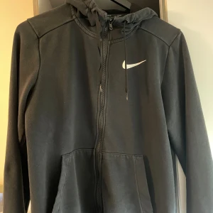Svart Nike hoodie med dragkedja - Svart hoodie från Nike med vit logga på bröstet. Tröjan har huva med snörning, hel dragkedja och två stora fickor framtill. Perfekt för chill eller träning, klassisk och enkel design i mjukt material.