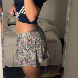 Blommig kortkjol i pastellfärger från H&M - Supersöt kortkjol med blommigt mönster lilla, blått, gult och svart. Kjolen har en flowy passform och resår i midjan för extra komfort. Kjolen har även två fickor på var sin sida som man inte ser så mycket på bilden för dem smälter värdligt bra in. Storlek XS men funkar lika mycket på S