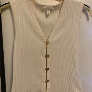 Ribbad beige linnetopp från H&M - Säljer en ribbad beige linnetopp från H&M med snygg v-ringning och dekorativa guldknappar framtill. Toppen är ärmlös och har en croppad passform som ger en fräsch och modern look. Perfekt att styla med jeans eller kjol för en trendig outfit.💕💕💕