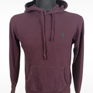 Vinröd hoodie från Ralph Lauren - Säljer en vinröd hoodie från Ralph Lauren med klassisk huva och snörning. Tröjan har en broderad grön logga på bröstet och en stor magficka framtill. Perfekt för dig som gillar en avslappnad och stilren look.