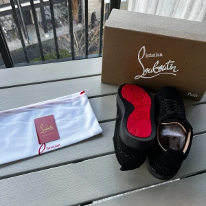 Christian Louboutin svarta sneakers mocka - Svarta sneakers från Christian Louboutin i lyxig mocka med ikonisk röd sula och broderad logga på hälen. Skorna har svarta snören, rund tå och platt sula. Kommer med originalkartong och dustbag. Perfekta för dig som vill sticka ut med exklusiv stil.
