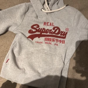 Grå Superdry hoodie med röd logga - Säljer en grå hoodie från Superdry med stor röd logga och japansk text framtill. Tröjan har huva med vita snören och en klassisk känguruficka. Perfekt för dig som gillar streetwear och vill ha en bekväm och snygg tröja.