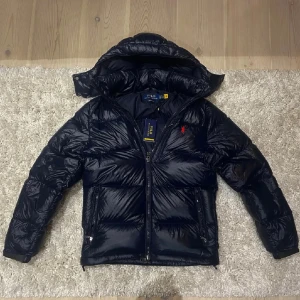 Mörkblå dunjacka från Polo Ralph Lauren - Snygg mörkblå dunjacka från Polo Ralph Lauren med huva och röd logga på bröstet. Jackan har glansig yta, två sidofickor med dragkedja och quiltad design. Perfekt för kalla dagar och har en modern, puffad look som sticker ut.