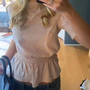 Ljusrosa blus med smock i midjan - Säljer en ljusrosa blus med rund hals och korta ärmar. Blusen har en smockad midja som ger en snygg volang nedtill. Materialet känns lätt och luftigt. Enkel och stilren design som är lätt att matcha.