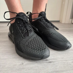 Svarta Nike Air Max 270 sneakers - Säljer ett par helsvarta Nike Air Max 270 sneakers med ventilerande mesh-ovandel, synlig luftenhet i hälen och klassisk snörning. Skorna har en sportig vibe och är riktigt bekväma med sin dämpade sula. Perfekta för dig som gillar streetstyle och vill ha en clean look.