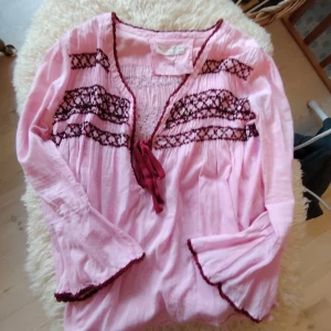 ODD MOLLY: Rosa Blus med Broderi - Supersöt rosa blus från Odd Molly med mörkbruna broderade detaljer och knytband framtill. Blusen har lång ärm med volangkant och v-ringad hals. Materialet är mjukt och lätt bomull, perfekt för vår och sommar. Broderade mönster ger en bohemisk vibe.