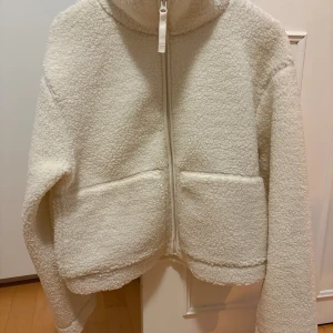 Vit teddyjacka i storlek M - Mysig vit teddyjacka med dragkedja framtill och stora fickor. Jackan har en hög krage och är tillverkad i fluffigt fleece-material som håller dig varm. Perfekt för kyliga dagar.