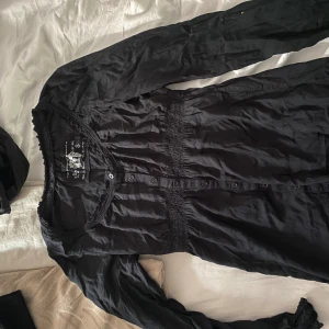 Svart långärmad klänning Odd Molly S - Svart långärmad klänning från Odd Molly i storlek S. Klänningen har v-ringad hals med spetsdetaljer, knappar framtill och smock vid midjan för en snygg passform. Materialet är mjukt och lätt, perfekt för dig som gillar stilrena och feminina plagg.