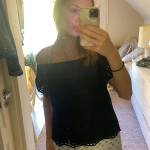 Svart offshoulder blus med broderi - Svart offshoulder blus i bomull med broderade detaljer och hålmönster längs ärmar och nederkant. Blusen har korta ärmar och en luftig passform, perfekt för varma dagar. Snygg och enkel att styla med jeans eller kjol.