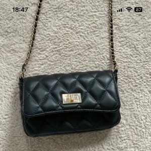 Svart quiltad axelväska med kedja - Snygg svart axelväska med quiltat mönster och guldfärgad kedja som axelrem. Väskan har ett stilrent lås framtill och insidan har ett fack med dragkedja. Perfekt storlek för det viktigaste och ger en cool vibe till din outfit.