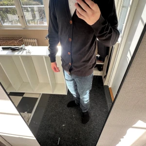 Mörkbrun stickad väst från Blend - Snygg mörkbrun stickad väst från Blend i storlek L. Västen har träfärgade knappar framtill, ribbade muddar och en liten Blend-lapp i skinn vid nederkanten. Perfekt för lager-på-lager och chill stil.