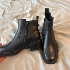 Svarta boots från Pace med klack - Snygga svarta boots från Pace i skinn med grov platt sula och låg klack. Modellen har rundad tå, dragkedja på sidan och robust design som ger en cool vibe. Perfekt för dig som gillar stilrena och tidlösa skor med edge.