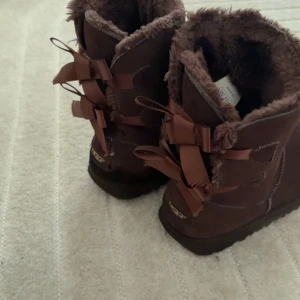 Bruna UGG boots med rosetter - Säljer ett par bruna UGG boots med fluffigt foder och dekorativa rosetter baktill. Skorna har platt sula och är tillverkade i mocka med mjuk päls på insidan. Perfekta för vintern! 