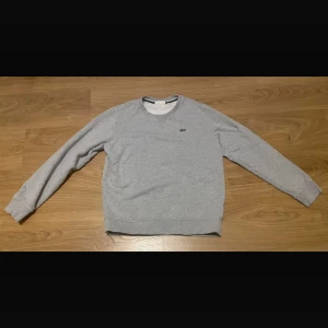 Grå sweatshirt från Lacoste - Snygg grå sweatshirt från Lacoste med klassisk rund hals och det ikoniska krokodilmärket broderat på bröstet. Tröjan har långa ärmar och ribbade muddar vid ärmslut och nederkant. Perfekt för en chill och stilren look.