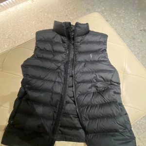 Svart dunväst från Polo Ralph Lauren - Pris går att diskutera vid snabb affär. Snygg svart dunväst från Polo Ralph Lauren med hög krage och diskret logga på bröstet. Västen har quiltad design, dragkedja framtill och två fickor. Perfekt för lager-på-lager och ger en clean look till din outfit. 