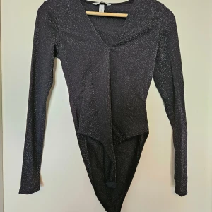 Svart glittrig body från H&M - Svart glittrig body från H&M med v-ringning och långa ärmar. Snyggt figurnära passform och tryckknappar i grenen. Perfekt för dig som vill sticka ut med lite extra glitter och glam.