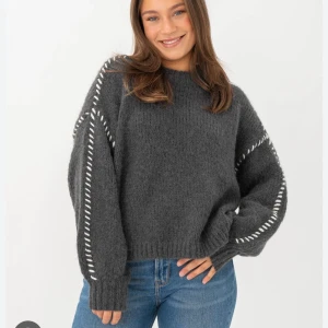 Mörkgrå stickad tröja med vita detaljer - vero Moda - Mysig mörkgrå stickad tröja med oversized passform och långa ärmar. Tröjan har vita dekorativa sömmar längs ärmarna och ribbade muddar vid ärmslut och nederkant. 