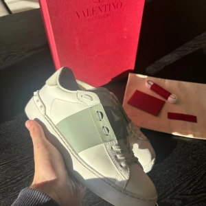 Valentino Garavani Open sneakers - Säljer ett par Valentino Garavani Open sneakers i vitt skinn med bred grön lackad rem över mitten. Klassisk rund tå, platt sula och ikoniska nitar baktill. Kommer med originalkartong, extra skosnören och dustbag. Perfekta för dig som gillar lyxiga detaljer.