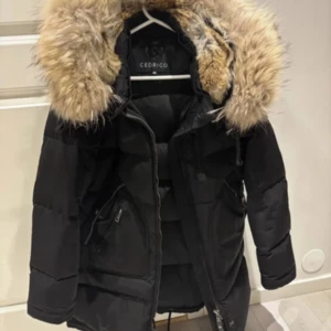 Svart pufferjacka med päls från Cedrico - Säljer en svart pufferjacka från Cedrico med stor fluffig päls på huvan. Jackan har dragkedja, långa ärmar och praktiska fickor med zip. Perfekt för dig som vill ha en varm och snygg vinterlook med statement-huva.
