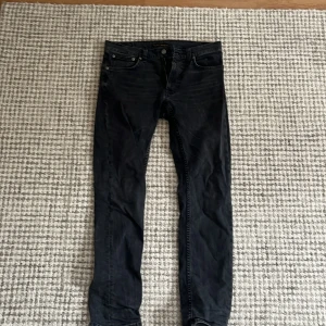 Svarta jeans från Nudie Jeans, 30/32 - Svarta jeans från Nudie Jeans med klassisk femficksdesign och diskret broderad bakficka. Jeansen har soft passform och är tillverkade i mjukt denimtyg. Perfekta för dig som gillar stilrena och tidlösa byxor. Måtten finns W30 och M32