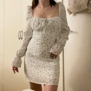 Vit blommig kortklänning med puffärm - Supersöt vit kortklänning med små blå blommor, långa puffärmar och fyrkantig urringning. Klänningen har knytband framtill och figursydd. Storlek xs och är ursprungligen från H&M. Dragkedjan är lite kärv men var det när jag köpte den💙🦋