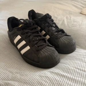 Adidas Superstar svarta sneakers - Klassiska Adidas Superstar sneakers. Super snygga men tyvärr så står dem bara i garderoben. Inte riktigt min stil längre därav säljer jag dem. Inga hål eller skador på dem förutom lite slitet vid hälarna. Mestadels damminga nu men annars unga märkbara fläckar på dem.