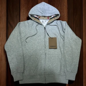 Grå hoodie från Burberry med dragkedja - Snygg grå hoodie från Burberry i 100% bomull med klassiskt rutigt foder i huvan. Tröjan har dragkedja, snörning i huvan och broderad Burberry-logga på bröstet. Perfekt för dig som gillar stilrena och exklusiva plagg.