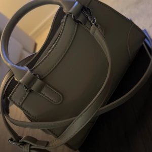 Beige handväska med dragkedja  - Snygg beige handväska i skinnimitation med både handtag och axelrem. Väskan har dekorativa detaljer på sidorna och en praktisk dragkedja framtill. Perfekt storlek för vardagen. Väskan är från Gina Tricot. Mycket bra skicka då jag använt den en gång. 