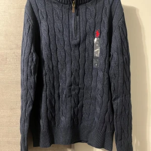 Polo Ralph Lauren half zip - Orginal pris: 2113kr, endast för 899kr hos Luxe Vibes!Kabelstickad marinblå tröja från Polo Ralph Lauren med halv dragkedja och röd broderad logga på bröstet. Tröjan har lång ärm, ribbade muddar och är tillverkad i mjuk bomull. Perfekt för lager-på-lager och snygg till jeans.