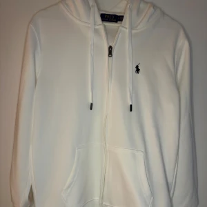 Vit hoodie från Polo Ralph Lauren - Säljer en stilren vit hoodie från Polo Ralph Lauren med dragkedja, huva och snörning. Tröjan har broderad logga på bröstet och känguruficka framtill. Tillverkad i 100% bomull för en skön och mjuk känsla. Perfekt för en clean och klassisk look.