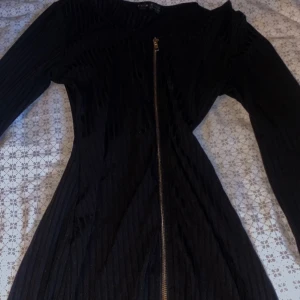 Svart långärmad klänning med dragkedja - Säljer en svart långärmad klänning med snygga ribbade detaljer och en synlig dragkedja framtill. Klänningen har en figurnära passform och är tillverkad i ett mjukt material som ger en cool och modern look. Perfekt för dig som gillar stilrena och edgy plagg.