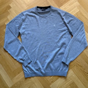 Grå stickad tröja Tiger of Sweden - Snygg grå stickad tröja från Tiger of Sweden i italienskt garn. Klassisk rund halsringning, diskret broderad logga på bröstet och ribbade muddar. Perfekt för dig som gillar stilren och enkel design.