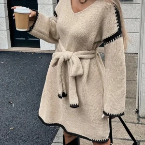 Beige stickad klänning med bälte - Supermysig beige stickad klänning med svarta detaljer vid ärmslut, axlar och nederkant. Klänningen har v-ringning, långa ärmar och ett matchande knytbälte i midjan. Perfekt för dig som gillar en chill och trendig look.