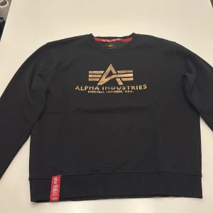 Svart sweatshirt Alpha Industries - Svart sweatshirt från Alpha Industries med guldig logga och text framtill. Tröjan är en fin tröja som man kan ha på sig när man vill. Snygg detalj med röd tag vid nederkanten. Tröjan är i ett väldigt bra skick då den är max använd 1-2 gånger. Tröjan har storleken M men passar folk som har storleken S. Pris kan diskuteras.