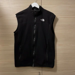 Svart fleeceväst från The North Face - Svart fleeceväst från The North Face i storlek M men sitter mer som en S. Västen har hög krage, hel dragkedja framtill och två fickor med dragkedja. Perfekt för lager-på-lager och outdoor-aktiviteter. Klassisk och stilren design med logga på bröstet.