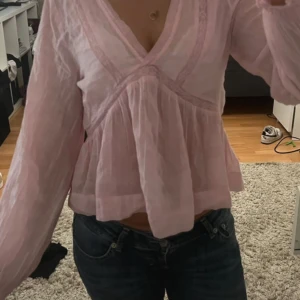 Ljusrosa blus med v-ringning - Säljer en ljusrosa blus med v-ringad hals och långärmade, lätt puffiga ärmar. Blusen har en volang nedtill och är tillverkad i ett tunt, luftigt material som ger en flowy känsla. Perfekt för dig som gillar romantisk och feminin stil.
