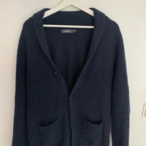 Mörkblå kofta från Dressmann - Säljer en mörkblå, ribbstickad kofta från Dressmann med två stora fickor framtill och knäppning med tre knappar. Koftan har en bred sjalkrage och är långärmad, perfekt för lager-på-lager-stil under kyliga dagar.