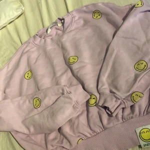 Ljuslila sweatshirt med smileys H&M - Superfin ljuslila sweatshirt från H&M med glada gula smiley-mönster över hela tröjan. Mjuk bomullskänsla, rund hals och ribbade muddar vid ärmslut och midja. Perfekt för dig som gillar färg och roliga prints!