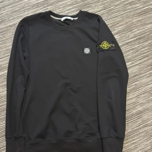 Svart sweatshirt från Stone Island - Svart långärmad sweatshirt från Stone Island med klassisk rund hals och ikonisk logotyp-patch på vänster ärm. Tröjan har ribbade muddar och är tillverkad i mjuk bomull för en skön och stilren look.