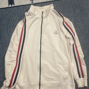 Vit Moncler ziptröja med ränder - Snygg vit ziptröja från Moncler med högt krage och dragkedja framtill. Tröjan har röda och marinblå ränder längs ärmarna och Moncler-logga på bröstet. Tillverkad i mjuk bomull, perfekt för en sportig och clean look.